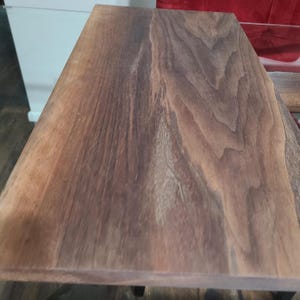 May include: Close-up of a rectangular wooden table top. The wood grain is visible, with a mix of light and dark brown tones. The edges of the table are smooth and finished. The table appears to be new and unused.