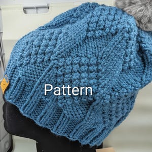 May include: A blue knit hat with a textured pattern and a gray faux fur pom-pom. The hat is displayed on a black mannequin head. The word "Pattern" is visible on the hat.