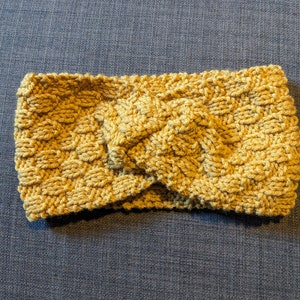 May include: Mustard yellow knitted headband with a textured, woven design. The headband is made of a soft, warm material and features a twisted knot detail at the front. This accessory is perfect for keeping hair out of the face.