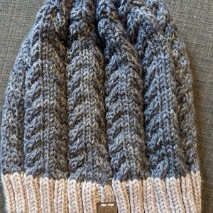May include: A gray and white knitted beanie with a cable knit pattern. The beanie has a tan colored brim and a brown leather tag with a logo on it.