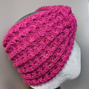 以下が含まれることがあります： ピンクの編み地のヘッドバンドで、テクスチャのあるパターンが施されています。ヘッドバンドはグレーのマネキンヘッドに表示されています。