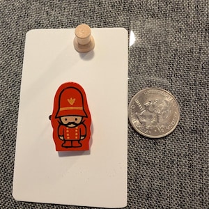 May include: A red and black wooden pin of a cartoon soldier with a gold emblem on a white card. A wooden push pin and a US quarter are also in the image. The background is a gray textured fabric.