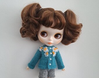 May include: A doll with brown hair styled in pigtails, wearing a teal knitted sweater with white and yellow daisy accents and pearl-like buttons. The doll is also wearing gray knitted shorts.