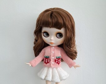May include: A doll with brown hair and large eyes wearing a pink cardigan with strawberry designs and a white skirt. The cardigan has pearl-like buttons. The doll is standing in front of a white background.
