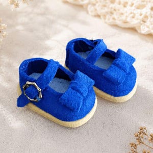 May include: A pair of miniature doll shoes in a vibrant royal blue color. These shoes feature a rounded toe, a decorative bow on the front, a single strap across the top, and a small metallic buckle. The soles are a light beige, flat material.