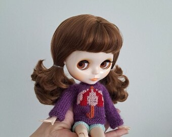 May include: A doll with brown hair styled in pigtails, wearing a purple sweater with a red and white umbrella design. The doll also has light blue shorts. The doll has large brown eyes and a fair complexion.