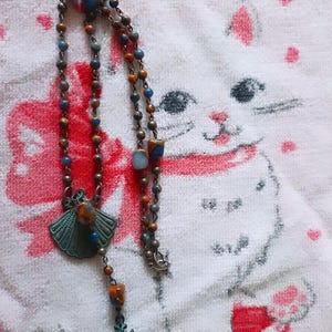 May include: A beaded necklace with blue, orange, and brown beads. The necklace has a decorative pendant and a cross charm. The background is a white towel with a cartoon cat design.