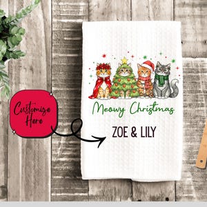 May include: White Christmas-themed kitchen towel featuring four cartoon cats dressed in festive attire. The towel reads "Meowy Christmas" and includes the names "Zoe & Lily". A wooden spoon rests on the side.