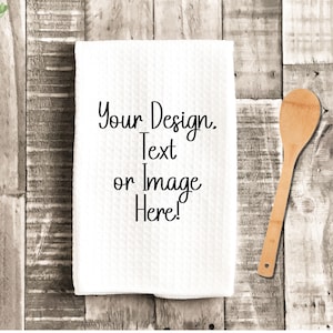 May include: White kitchen towel with a textured pattern, featuring the text "Your Design. Text or Image Here!". A wooden spoon rests on a rustic wooden surface, with a small potted plant in the upper left corner.