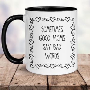 May include: White ceramic coffee mug with a black rim and handle. The mug has a black outline of a heart border with the text "Sometimes good moms say bad words" inside.