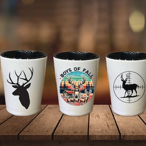May include: Three white ceramic shot glasses with black rims. The first glass has a silhouette of a deer head. The second glass has a deer silhouette with the text "Boys of Fall". The third glass has a deer silhouette in a target scope.