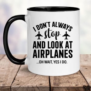 May include: White ceramic mug with a black handle and rim. The mug features the text "I DON'T ALWAYS stop AND LOOK AT AIRPLANES ...OH WAIT, YES I DO." in black, with airplane graphics. The mug is on a wooden surface.