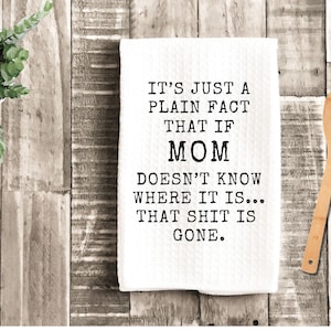 May include: A white kitchen towel with the text "IT'S JUST A PLAIN FACT THAT IF MOM DOESN'T KNOW WHERE IT IS... THAT SHIT IS GONE." A wooden spoon and a potted plant are also in the image.