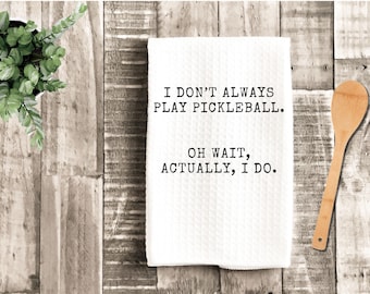 May include: White kitchen towel with the text "I DON'T ALWAYS PLAY PICKLEBALL. OH WAIT, ACTUALLY, I DO." A wooden spoon and a potted plant are also in the image. The towel is on a wooden surface.