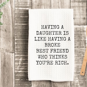 K&ouml;nnte beinhalten: Wei&szlig;es K&uuml;chentuch mit dem Text "HAVING A DAUGHTER IS LIKE HAVING A BROKE BEST FRIEND WHO THINKS YOU'RE RICH." Ein Holzl&ouml;ffel liegt neben dem Tuch auf einer Holzoberfl&auml;che.