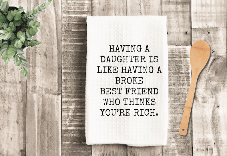 K&ouml;nnte beinhalten: Wei&szlig;es K&uuml;chentuch mit dem Text "HAVING A DAUGHTER IS LIKE HAVING A BROKE BEST FRIEND WHO THINKS YOU'RE RICH." Ein Holzl&ouml;ffel liegt neben dem Tuch auf einer Holzoberfl&auml;che.