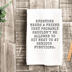 May include: White kitchen towel with the text "EVERYONE NEEDS A FRIEND THEY PROBABLY SHOULDN'T BE ALLOWED TO SIT NEXT TO AT SERIOUS FUNCTIONS." A wooden spoon and a small potted plant are also visible.