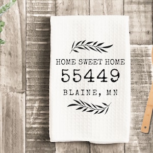 May include: White kitchen towel with the text "HOME SWEET HOME" and the numbers "55449" and the city "BLAINE, MN" in black. The towel has a textured pattern and is decorated with black leaf sprigs. A wooden spoon rests on a wooden surface.