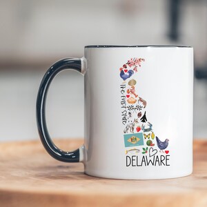 May include: White ceramic mug with a black handle and rim. The mug features a colorful illustration of the state of Delaware, including the text "DELAWARE" and "The First State". The design includes various state symbols.