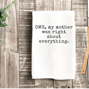 May include: White kitchen towel with the text "OMG, my mother was right about everything." A wooden spoon rests on a wooden surface next to the towel. A small potted plant is in the upper left corner.