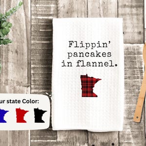 May include: White kitchen towel with the text "Flippin' pancakes in flannel." A red and black plaid outline of the state of Minnesota is below the text. A wooden spoon and a small potted plant are also in the image.
