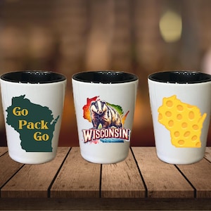 May include: Three white shot glasses with black rims. The glasses feature different Wisconsin-themed designs: a green outline of the state with the text "Go Pack Go", a badger with the text "Wisconsin", and a yellow outline of the state with a cheese pattern.