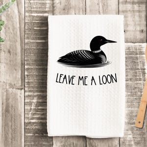 May include: White kitchen towel with a black graphic of a loon floating on water. The text "LEAVE ME A LOON" is printed below the loon. A wooden spoon rests on a wooden surface.
