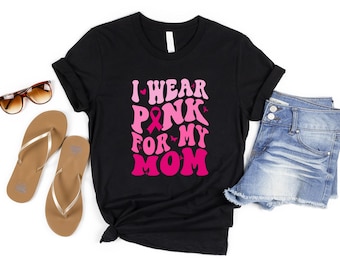 May include: Black t-shirt with the text "I Wear Pink For My Mom" in pink. The shirt is styled with sunglasses, tan flip-flops, and denim shorts. The design includes a pink ribbon, symbolizing breast cancer awareness.