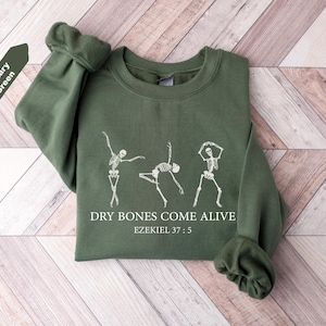 May include: Military green sweatshirt featuring a white graphic of three dancing skeletons. The text "DRY BONES COME ALIVE" and "Ezekiel 37:5" are printed below the skeletons. The sweatshirt has a crew neck and long sleeves.