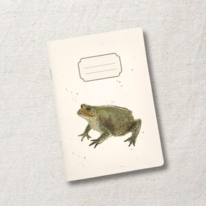 Könnte beinhalten: Ein cremefarbenes Notizbuch mit einer detaillierten Illustration einer grünen und braunen Kröte. Das Notizbuch hat oben ein leeres Etikett und kleine graue Sprenkel um die Kröte. Der Einband ist leicht angewinkelt und zeigt die Ecke.