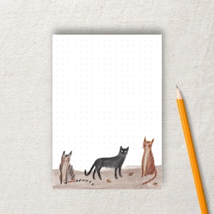 Könnte beinhalten: Ein weißer Notizblock mit einem gepunkteten Rastermuster und einem Bleistift. Der untere Teil des Notizblocks zeigt Illustrationen von drei Katzen: einer Tabbykatze, einer schwarzen Katze und einer roten Katze. Die Katzen stehen auf einer braunen Oberfläche.