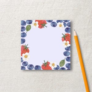 Könnte beinhalten: Ein quadratischer Notizblock mit hellviolettem Hintergrund und einem Rand aus Aquarellillustrationen von roten Erdbeeren, weißen Erdbeerblüten, grünen Blättern und dunkelblauen Heidelbeeren, dazu ein gelber Holzbleistift.