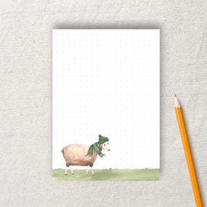 Könnte beinhalten: Ein Notizblock mit einer Aquarellillustration eines Schafs mit grüner Mütze und Schal. Der Notizblock hat ein gepunktetes Rastermuster und ein Bleistift liegt daneben.