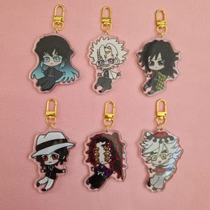 Könnte beinhalten: Sechs Schlüsselanhänger aus Acryl mit Anime-Charakteren und goldfarbenen Verschlüssen. Die Charaktere haben verschiedene Haarfarben und Outfits mit detaillierten Gesichtsausdrücken. Die Schlüsselanhänger befinden sich auf einem rosa Hintergrund.
