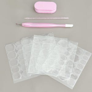 May include: A nail care kit featuring a pink buffer, a clear cuticle pusher, a pink and white cuticle tool, and several sheets of clear nail adhesive tabs. The tools are arranged on a grey surface.