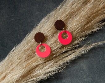 May include: A pair of earrings featuring a dark brown wooden circle at the top, connected to a vibrant neon pink, half-circle pendant. The earrings are displayed against a backdrop of dried beige grass and a dark gray surface.