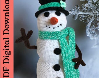 May include: A crocheted snowman with a black top hat, a green scarf, and black buttons. The snowman has a carrot nose and black eyes.