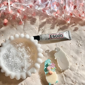 May include: A white ceramic dish filled with clear plastic heart-shaped charms. A tube of E6000 glue is next to the dish. A partially assembled gnome ornament with a turquoise hat and pink flowers is on a white surface.