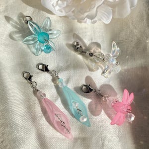 May include: A collection of five decorative charms with silver-tone clasps. The charms feature various designs, including a light blue flower, a clear iridescent fairy, and three teardrop-shaped charms in pink and light blue. Each charm has bead accents.