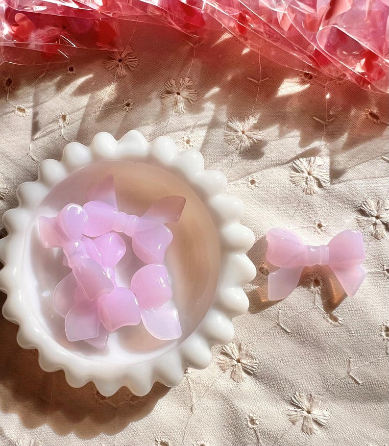 May include: A collection of translucent pink bow-shaped embellishments. Some bows are in a white, scalloped-edge dish, while others are scattered on a cream-colored, embroidered fabric. The bows have a glossy finish and are likely decorative.