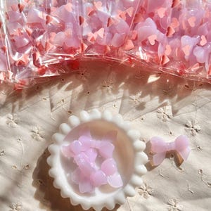 May include: A collection of pink bow-shaped embellishments, some packaged in clear plastic bags, others in a white, scalloped-edge dish. The bows are a translucent pink, and small pink heart-shaped confetti is scattered around.