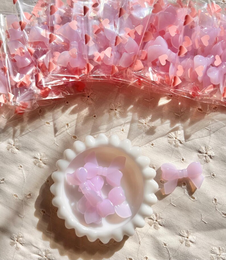 May include: A collection of pink bow-shaped embellishments, some packaged in clear plastic bags, others in a white, scalloped-edge dish. The bows are a translucent pink, and small pink heart-shaped confetti is scattered around.