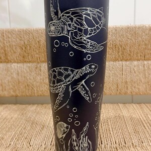 Peut inclure: Une gourde isotherme bleu marine avec un motif de tortue de mer argenté. La gourde a un couvercle à bouton-poussoir et une poignée. Le motif comprend deux tortues, des bulles, des algues et un poisson. La gourde est conçue pour garder les boissons froides.