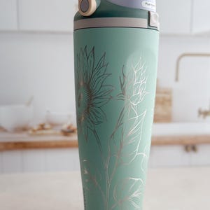 Peut inclure: Une gourde isotherme vert menthe avec une poignée et un couvercle beiges. La bouteille présente un motif floral argenté de tournesols et de feuilles. Le couvercle a un bouton-poussoir pour une ouverture facile. La bouteille est conçue pour garder les boissons froides pendant des heures.