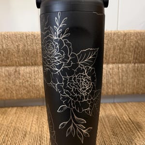 Peut inclure: Gourde isotherme noire avec un motif floral argenté. La gourde a une poignée et un couvercle noirs. Le motif floral comprend des roses et des feuilles. Conçue pour garder les boissons froides ou chaudes. Un accessoire élégant et pratique.