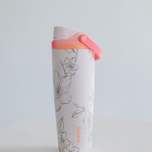 Peut inclure: Une gourde Owala blanche avec un motif floral. La bouteille a une poignée et un couvercle roses avec un accent orange. Le motif floral est en gris. La bouteille est sur une surface blanche.