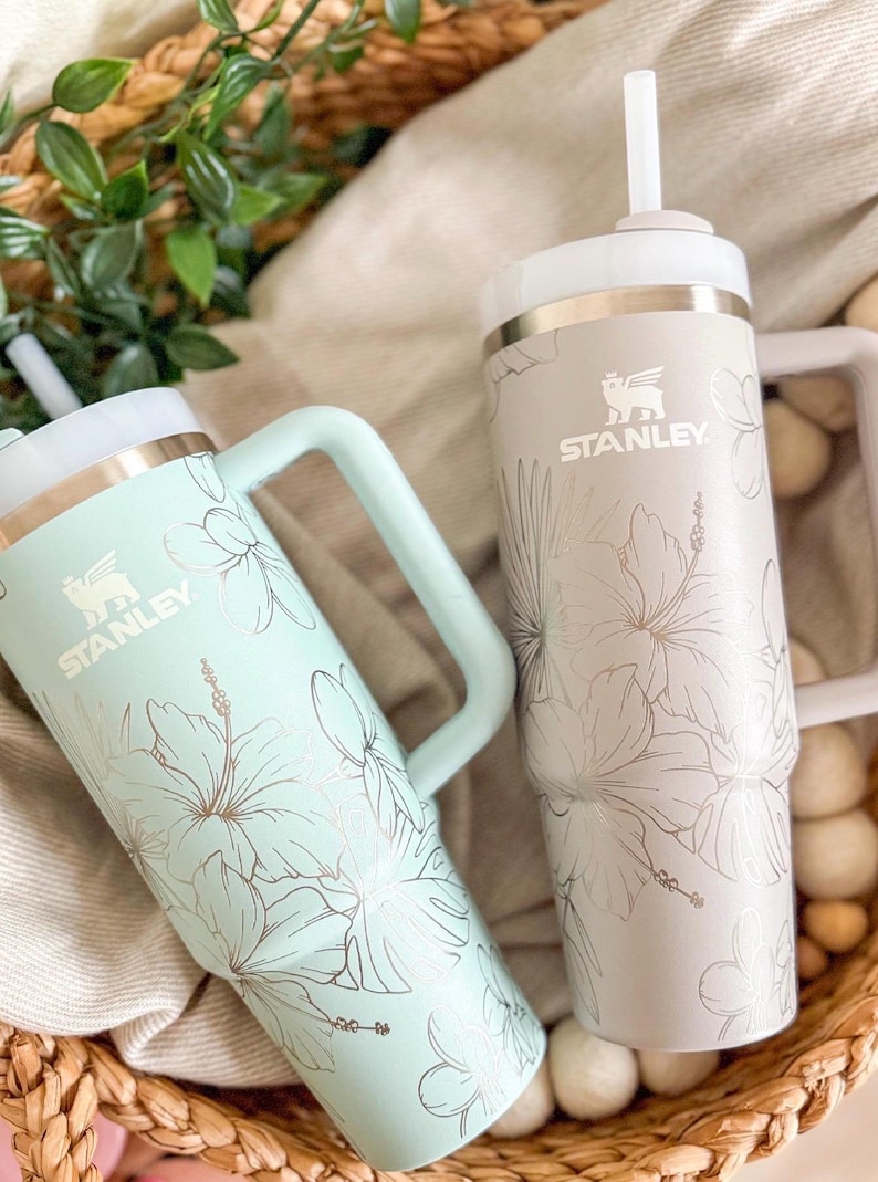 Pode incluir: Duas canecas Stanley com al&ccedil;as e canudos. Uma &eacute; verde menta, a outra bege. Ambas apresentam um design floral e o logotipo Stanley. Est&atilde;o em uma cesta de vime com vegeta&ccedil;&atilde;o.