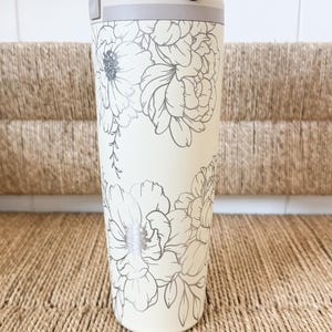 Peut inclure: Gourde crème avec un motif floral argenté. Elle possède une poignée rose clair et un couvercle blanc. Le motif floral présente de grandes fleurs de pivoines. La gourde est posée sur une surface tissée.