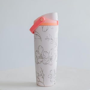 Peut inclure: Un gobelet blanc à motifs floraux avec une poignée et un couvercle roses. Le gobelet présente une bande d'accent rose clair et orange. Le motif floral est de couleur gris argenté. Le gobelet est sur une surface blanche.