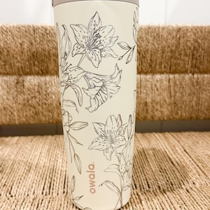 Peut inclure: Une gourde crème avec un motif floral et une poignée beige. La bouteille présente un motif répété de fleurs de lys argentées. La marque "owala" est imprimée sur le côté.